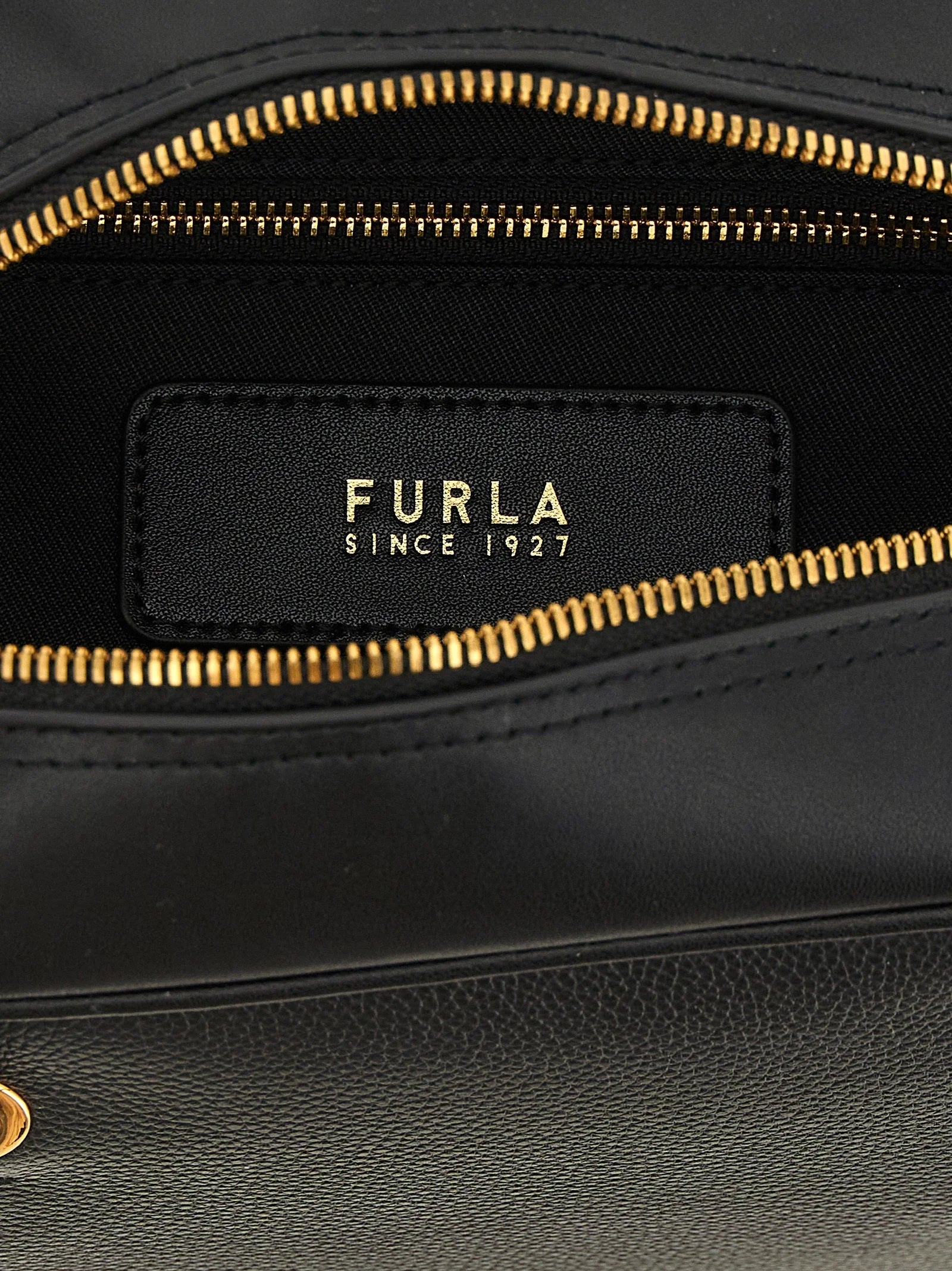 Detalhe close-up da mala Furla, mostrando a grão de couro de bezerro premium e o fecho metálico polido com o logótipo.